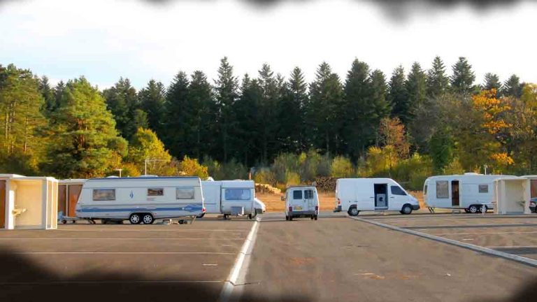 Photo d'une aire de grand passage avec plusieurs caravanes et fourgons stationnés, forêt en arrière-plan