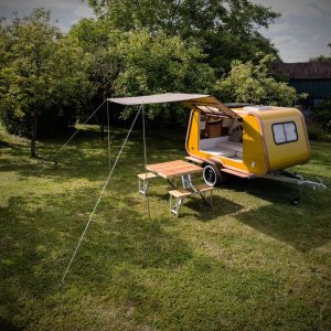 Mini-caravane Carapate de couleur jaune installée sur une pelouse arborée, avec la porte arrière ouverte, un auvent déployé et une table de camping avec deux bancs positionnée à l’extérieur pour un repas en plein air.