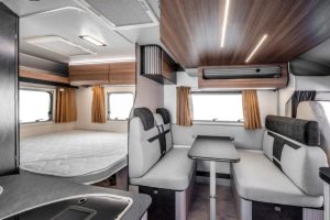 : Intérieur moderne d’un camping-car avec un espace nuit équipé d’un lit double à l’arrière, une salle à manger avec banquettes et table au centre, et un coin cuisine au premier plan. Mobilier en bois foncé et tissus gris
