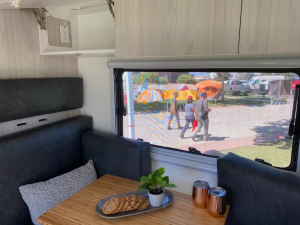 Espace repas confortable à l’intérieur d’un van, avec banquettes et petite table. À travers la fenêtre, on distingue une scène extérieure animée avec des tentes et des passants.