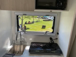 Vue intérieure d’un van aménagé moderne avec un coin cuisine équipé d’un évier et d’une plaque à induction. Par la fenêtre, on aperçoit un parc verdoyant