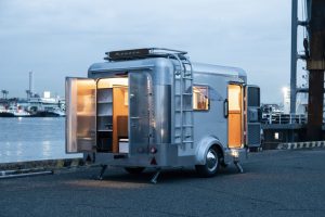 Mini caravane japonaise X-Cabin 300 en aluminium avec intérieur éclairé et portes ouvertes, au bord de l'eau au crépuscule