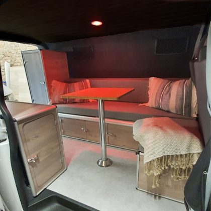 Espace de vie chaleureux dans un van équipé du kit Sunset Van avec banquette, coussins, table amovible et lumière d’ambiance rouge
