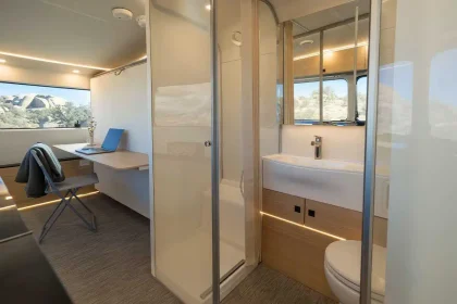 Salle de bains compacte et lumineuse dans une caravane moderne, avec douche vitrée, lavabo design et vue extérieure sur paysage désertique.