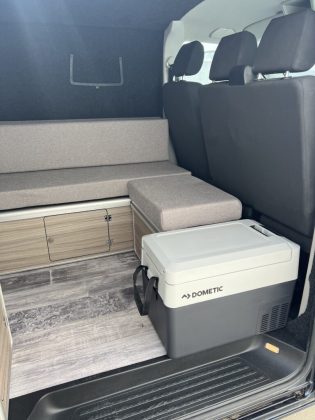 Vue intérieure d’un utilitaire aménagé avec le kit Sunset Van : banquette convertible, rangements en bois clair et glacière Dometic
